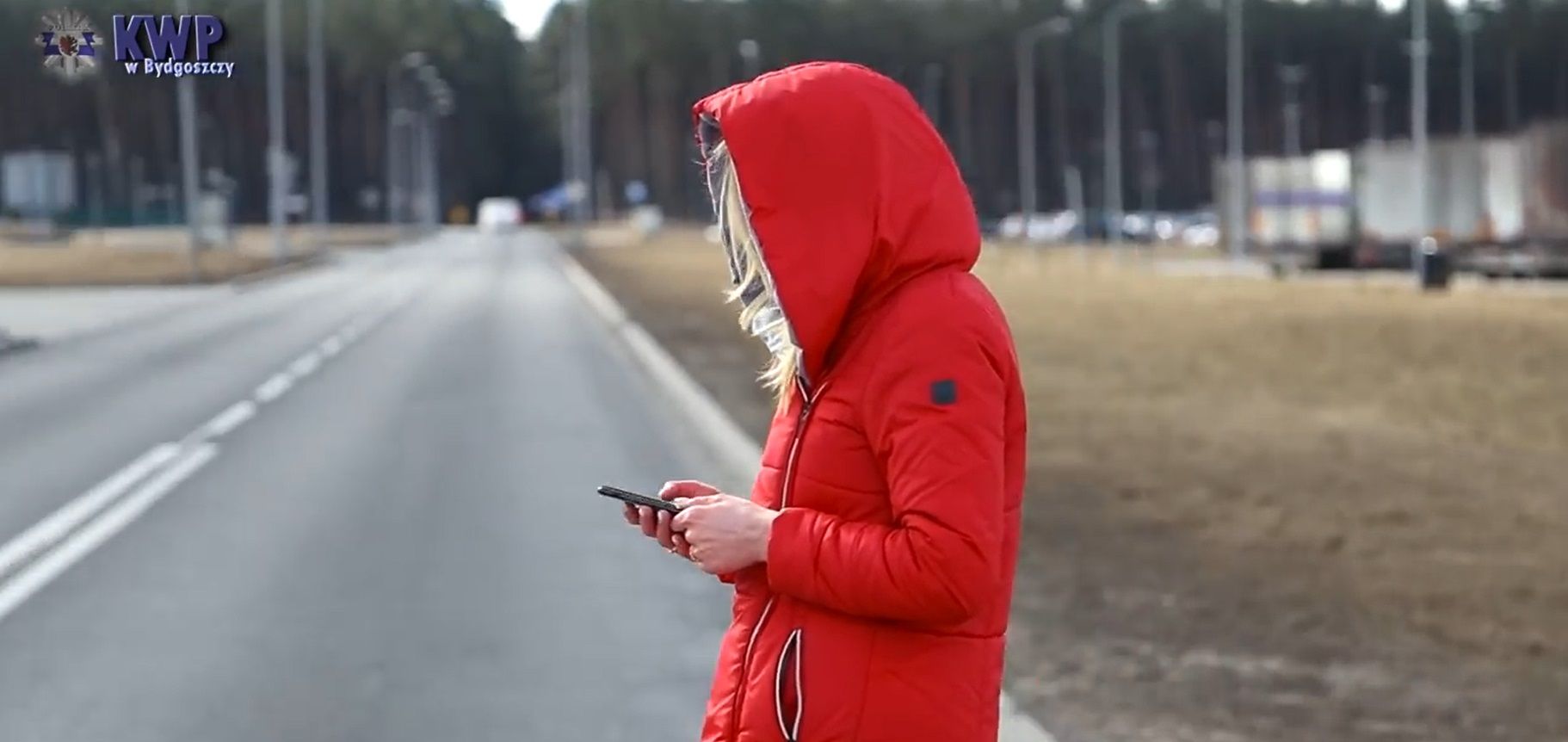 Kobieta z kapturem na głowie wpatrzona w smartfon podczas przechodzenia przez ulicę.