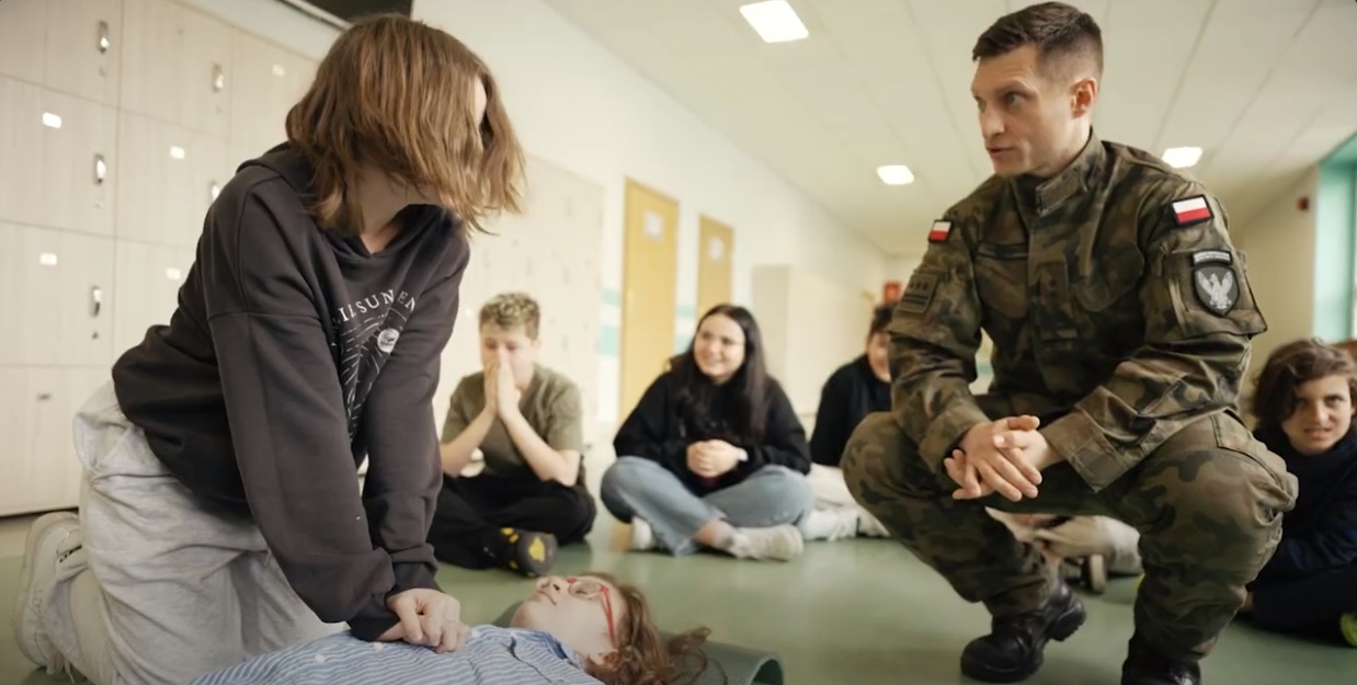 Nastolatka wykonuje masaż serca na zajęciach z pierwszej pomocy pod okiem żołnierza w mundurze.