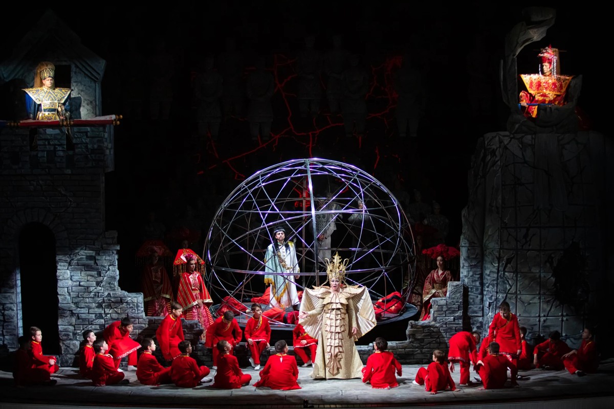przedstawienie operowe, kobieta w złotym stroju i koronie stoi przez siedzącymi wkoło chłopcami w czerwonych szatach, za nią dekoracje, ruiny zamku, kula i inne postaci