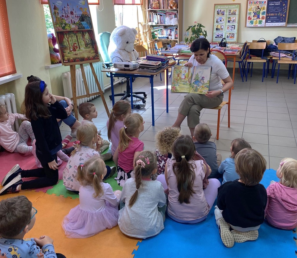 Kobieta prezentuje książkę małym dzieciom siedzącym na kolorowych matach.