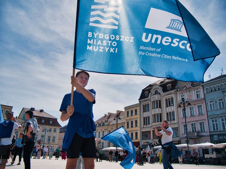 Na rynku nastolatek trzyma flagę z logo UNESCO i znakiem promocyjnym: Bydgoszcz - miasto muzyki.