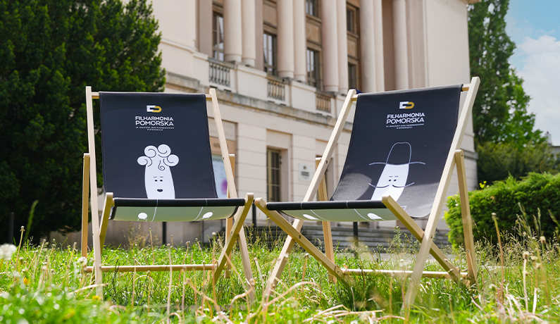 Dwa puste leżaki z logotypami Filharmonii Pomorskiej na trawniku przed budynkiem w stylu neoklasycystycznym.