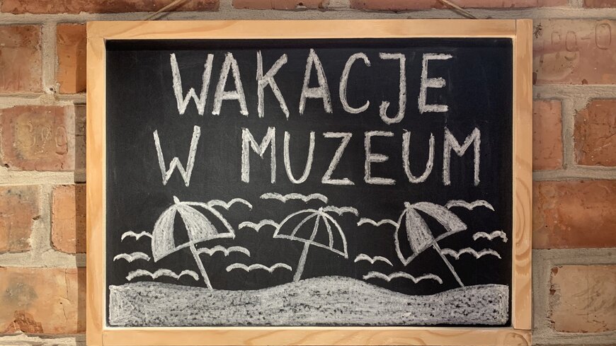 Grafika przedstawiająca w tle ceglaną ścianę, a na niej czarną tablicę szkolną z białym napisem wakacje w muzeum i plażowymi parasolami.