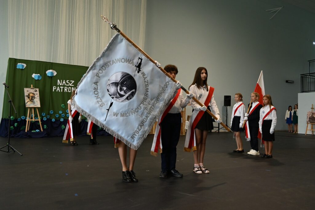 Uczniowie przepasani szarfami biało-czerwonymi trzymają sztandar szkolny podczas uroczystości.
