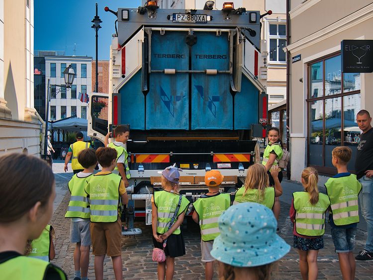 Na zdjęciu dzieci ubrane w kamizelki odblaskowe patrzące w stronę pojazdu do zbiórki odpadów komunalnych