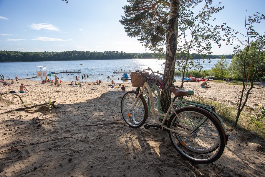 Na pierwszym planie dwa rowery oparte o drzewo. Ludzie siedzą i leżą na plaży nad jeziorem, w oddali las.