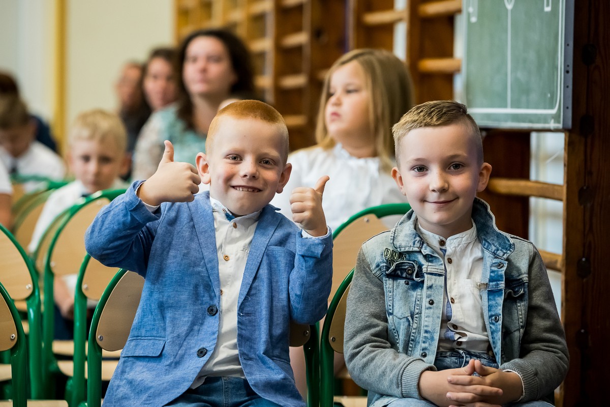 Uśmiechnięte dzieci siedzą na krzesłach w szkole, prawdopodobnie w większej sali. Na pierwszym planie dwóch chłopców, jeden z nich ma podniesione w górę oba kciuki.