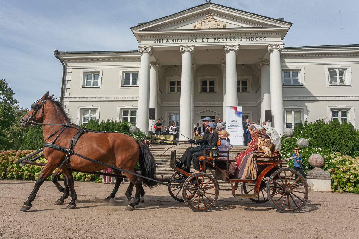 Powóz konny z pasażerami w strojach z XIX w. stoi przed budynkiem z kolumnami