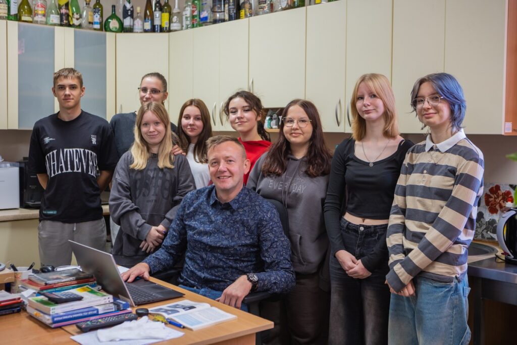 uśmiechnięty nauczyciel siedzi przy biurku w pracowni, za nim stoją uśmiechnięte uczennice i uczeń