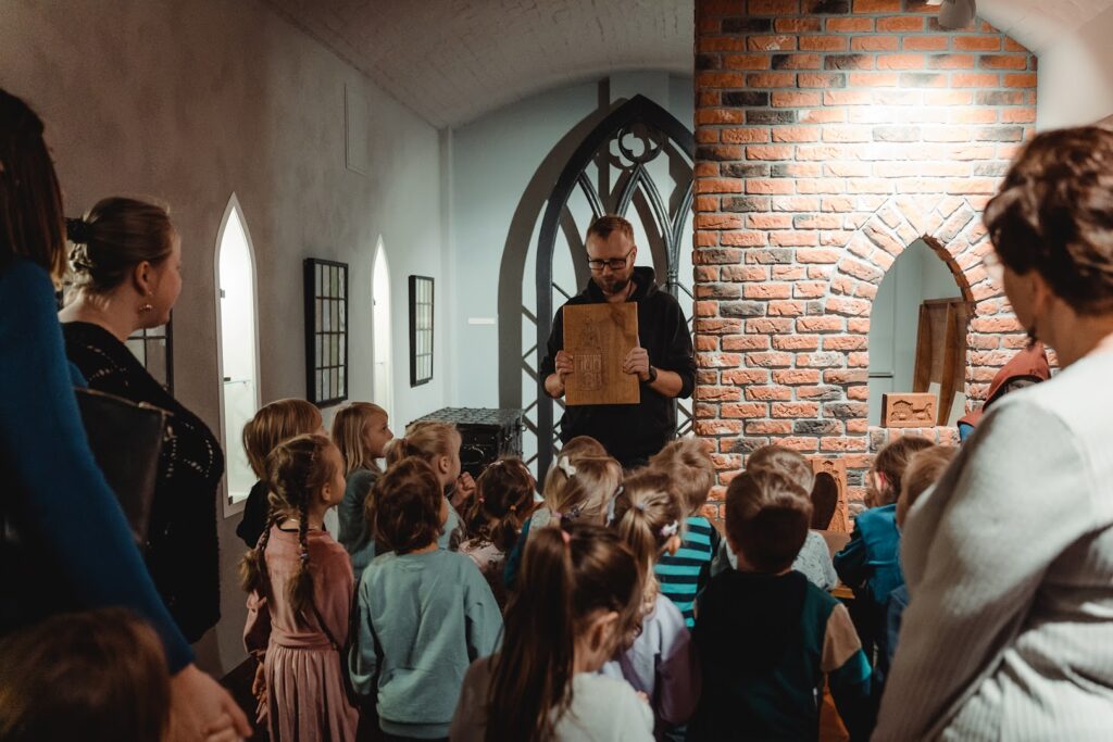 Mężczyzna z brodą, w okularach pokazuje grupie dzieci stary dokument