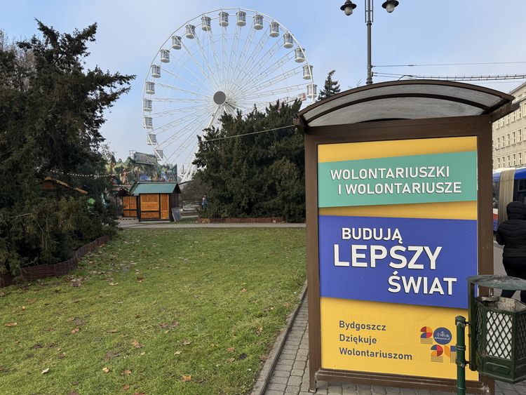 Na pierwszym planie plakat przy przystanku komunikacyjnym z napisem Wolontariuszki i wolontariusze budują lepszy świat. W tle diabelski młyn